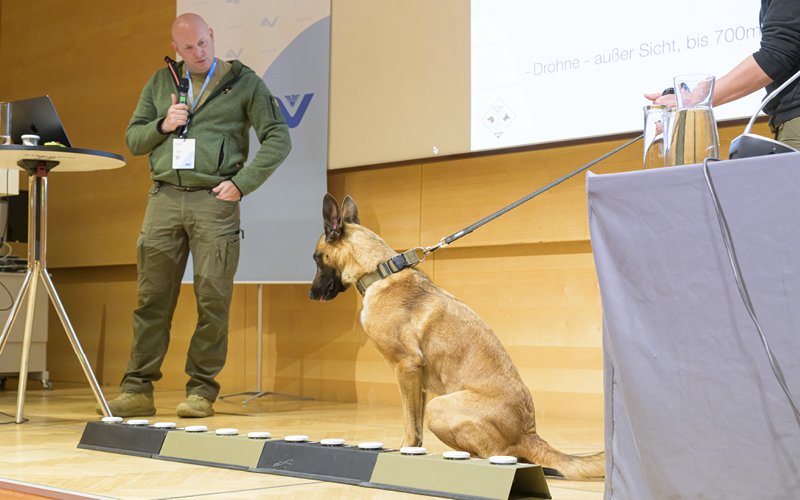 Mann mit Mikrofon hält einen Vortrag auf der Bühne neben einem Diensthund.