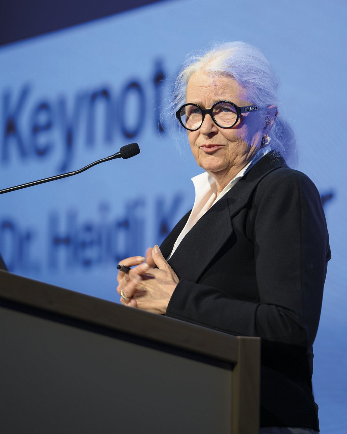 Dr.in Heidi Kastner spricht am Rednerpult vor blauem Hintergrund mit groß eingeblendetem Schriftzug; sie trägt eine schwarze Brille und einen dunklen Blazer.