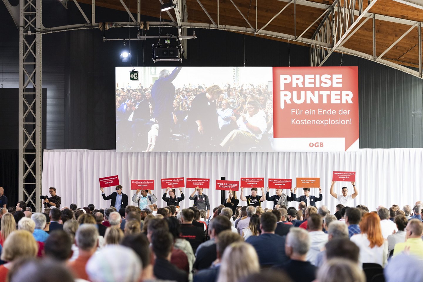 Menschenmenge vor einer Bühne mit jungen Menschen, die Tafeln mit ihren Forderungen hochhalten. Dahinter ein Screen mit Preise Runter Logo