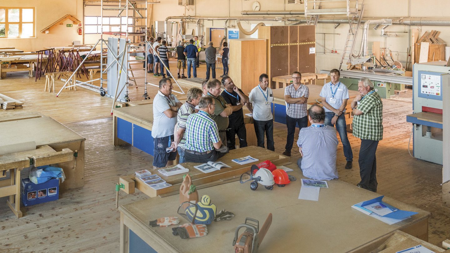 zwei Gruppen älterer Männer stehen in einer großen Halle eines holzverarbeitenden Betriebs besammen und besprechen sich