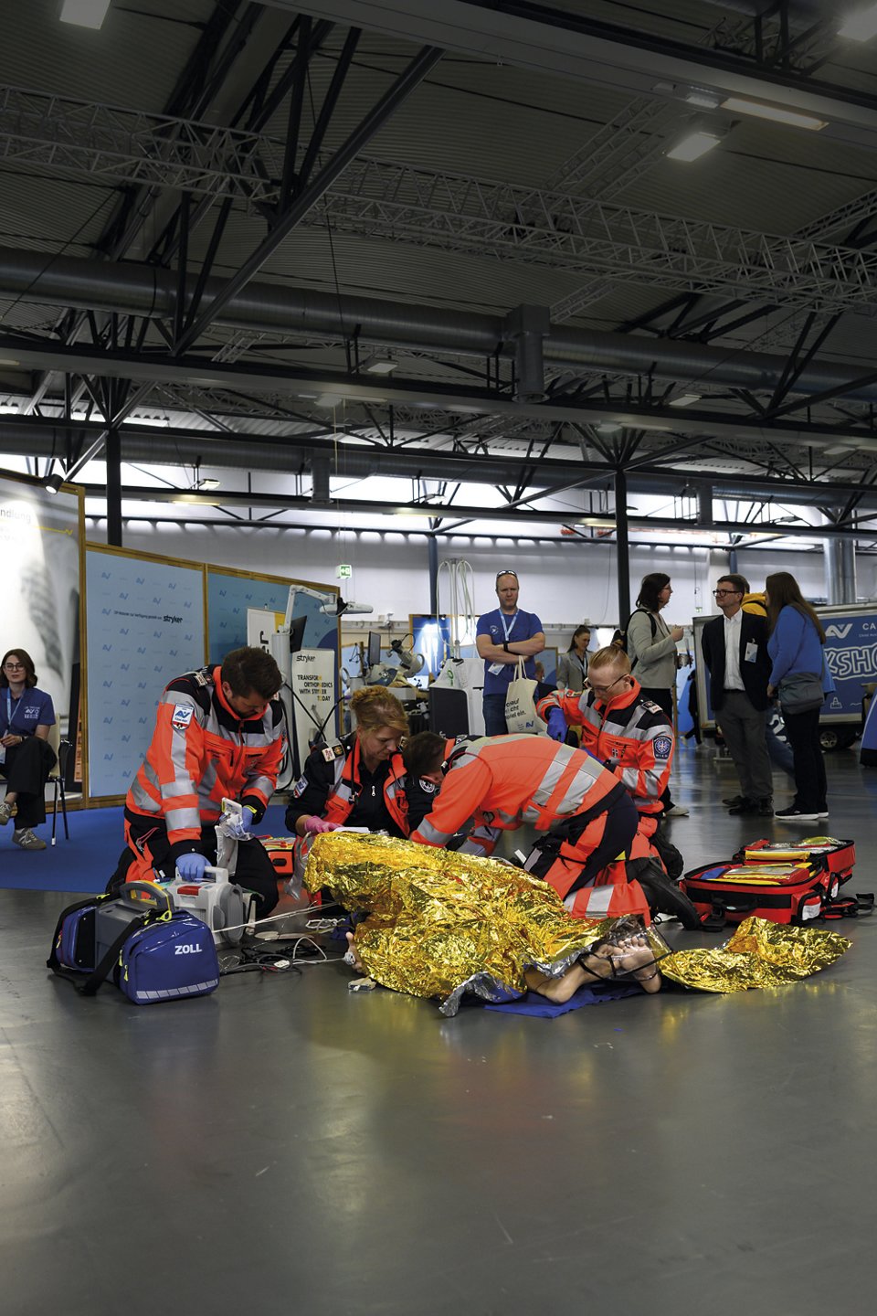 Sanitäter:innen in orangefarbener Einsatzkleidung leisten Erste Hilfe an einer Person mit Rettungsdecke auf dem Boden; medizinisches Equipment liegt bereit, mehrere Zuschauer beobachten die Szene.
