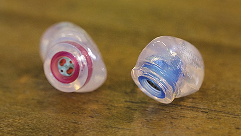 Zwei individuell angepasste Gehörschutz-Otoplastiken in Rot und Blau auf einem Holzuntergrund