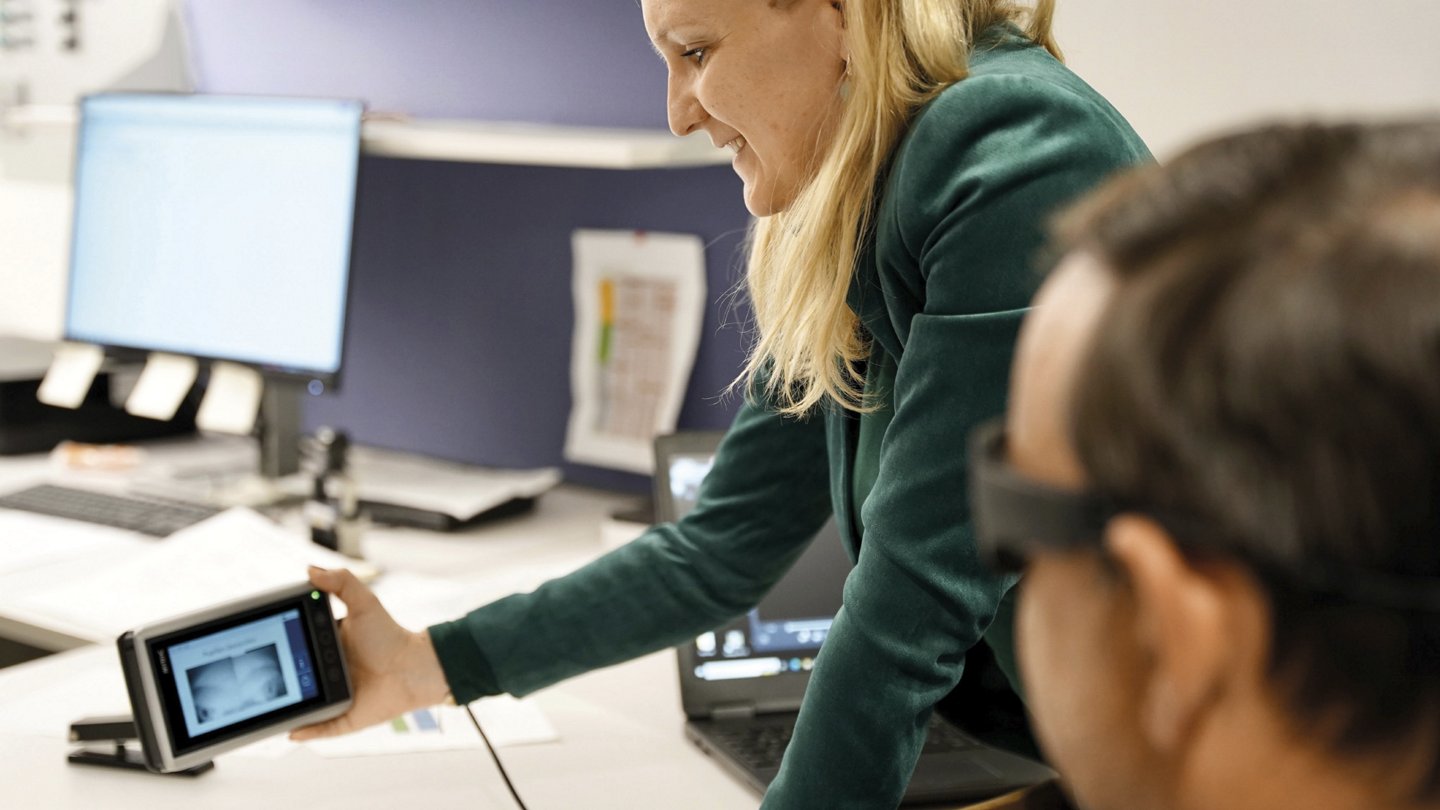 Eine Frau zeigt einem Mann, der eine spezielle Brille trägt, ein kleines elektronisches Gerät, vermutlich ein Bildschirm oder Monitor. Die beiden befinden sich in einem modernen Büro mit Computern und Dokumenten im Hintergrund.