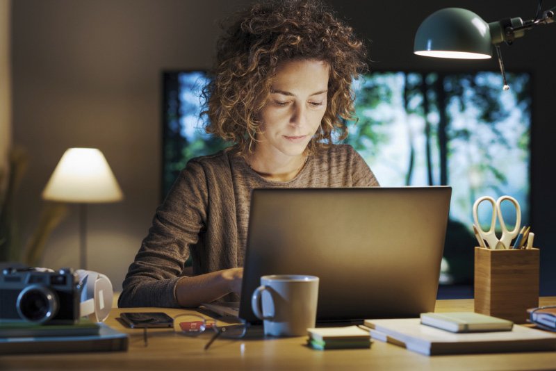 Eine Frau mit lockigem Haar arbeitet abends an einem Laptop, umgeben von Büromaterialien, mit einer Schreibtischlampe als Lichtquelle.