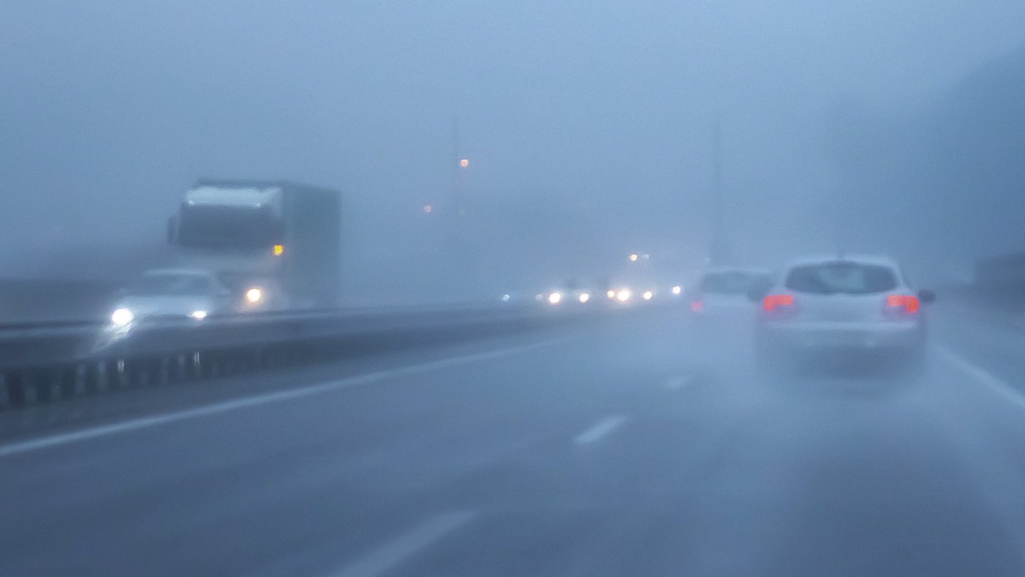 Autos und ein Lkw fahren auf einer Autobahn bei dichtem Nebel und nasser Fahrbahn. Die Scheinwerfer und Rücklichter sind eingeschaltet, um die Sichtbarkeit zu verbessern.