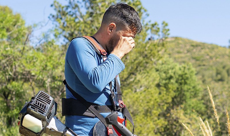Gartenarbeiter mit Freischneider hält sich erschöpft die Hand vors Gesicht –  symbolisiert Hitzestress und UV-Belastung bei Außenarbeit