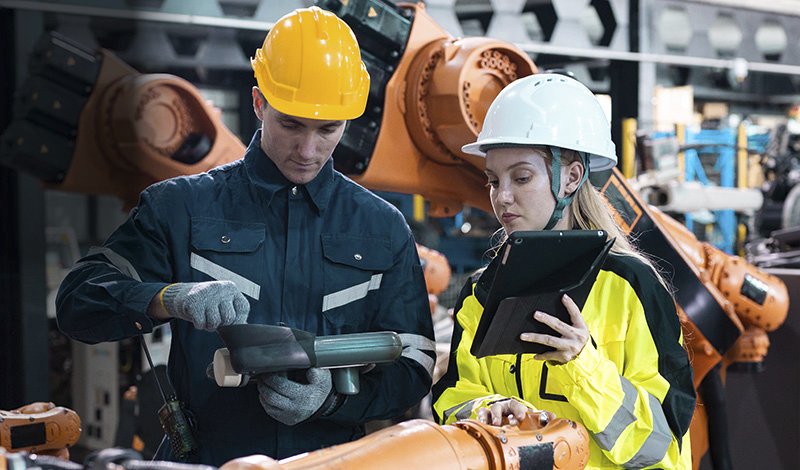 Zwei Beschäftigte mit Schutzhelmen arbeiten gemeinsam mit einem Industrieroboter – symbolisiert Mensch-Maschine-Kollaboration