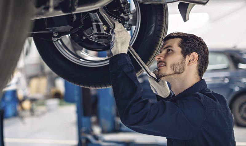 Ein Mechaniker in blauer Arbeitskleidung arbeitet unter einem angehobenen Auto in einer Werkstatt. Er trägt Handschuhe und benutzt einen Schraubenschlüssel, um an der Radaufhängung zu arbeiten.