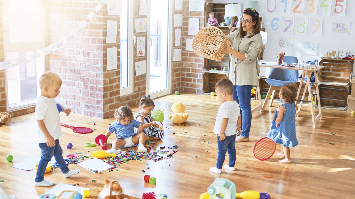 Eine Erzieherin steht in einem sonnendurchfluteten Kindergartenraum und hält einen leeren Korb in den Händen. Um sie herum spielen mehrere Kleinkinder mit Bauklötzen, Bällen und anderen Spielzeugen. Der Boden ist mit bunten Spielsachen bedeckt. An den Wänden hängen Kinderzeichnungen und Zahlen. Im Hintergrund steht ein Schreibtisch mit Stiften und Papier.