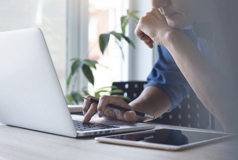 Eine Person sitzt an einem Schreibtisch und arbeitet an einem Laptop. In der Hand hält sie einen Stift, während ein Tablet auf dem Tisch liegt. Im Hintergrund sind grüne Pflanzen vor einem Fenster zu sehen.