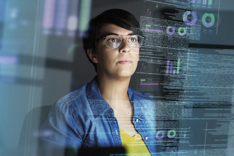 Bild einer jungen Frau mit Brille, die vor einem transparenten Bildschirm mit Datenvisualisierungen steht. Oben rechts ein Bereich mit einem unscharfen oder verpixelten Element.