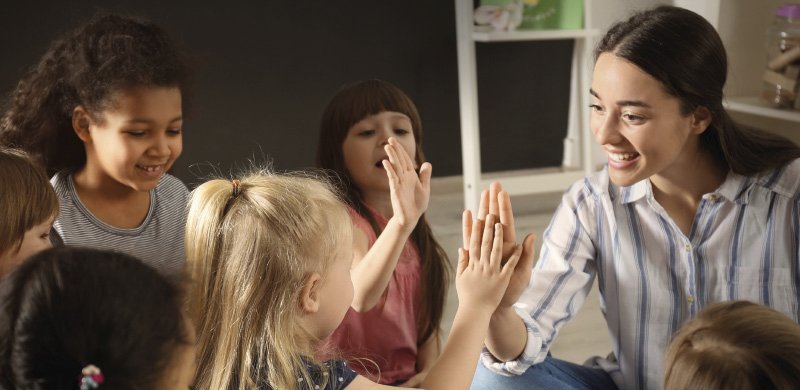 Eine junge Erzieherin mit langen dunklen Haaren und einem gestreiften Hemd gibt mehreren lachenden Kindern in einem Kindergarten High-Fives. Die Kinder, die unterschiedliche Haut- und Haarfarben haben, strahlen Freude und Begeisterung aus. Der Hintergrund zeigt ein Regal mit Spielzeug und Bastelmaterialien.