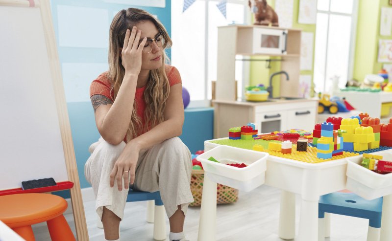 Eine junge Erzieherin mit Brille und langen, gewellten Haaren sitzt erschöpft in einem Kindergarten auf einem kleinen Stuhl. Sie stützt ihren Kopf auf eine Hand und wirkt müde. Neben ihr steht ein Tisch mit bunten Bauklötzen, im Hintergrund sind eine Spielküche und eine Maltafel zu sehen. Der Raum ist bunt gestaltet und für Kinder eingerichtet.