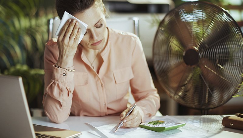 Frau sitzt erschöpft und gestresst vor einem Laptop in einem überhitzten Innenraum