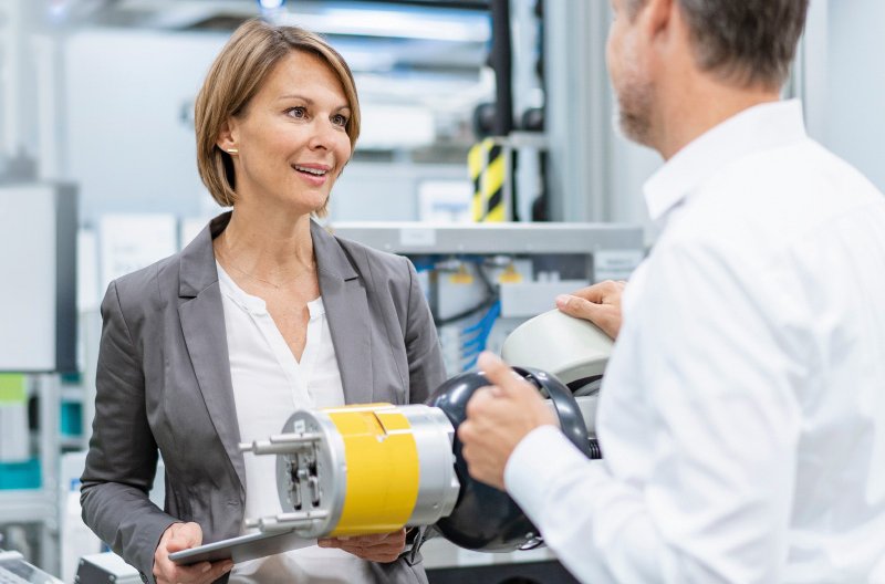 Eine Frau in einem grauen Blazer hält ein technisches Bauteil und spricht mit einem Mann in weißem Hemd in einer industriellen Umgebung.