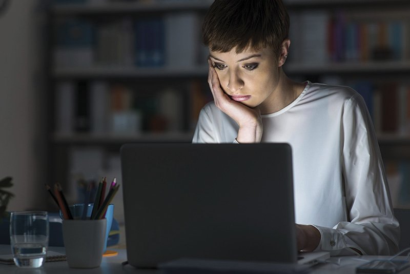 eine Frau sitzt im Licht ihres Notebooks und stützt ihren Kopf auf ihren Händen ab.