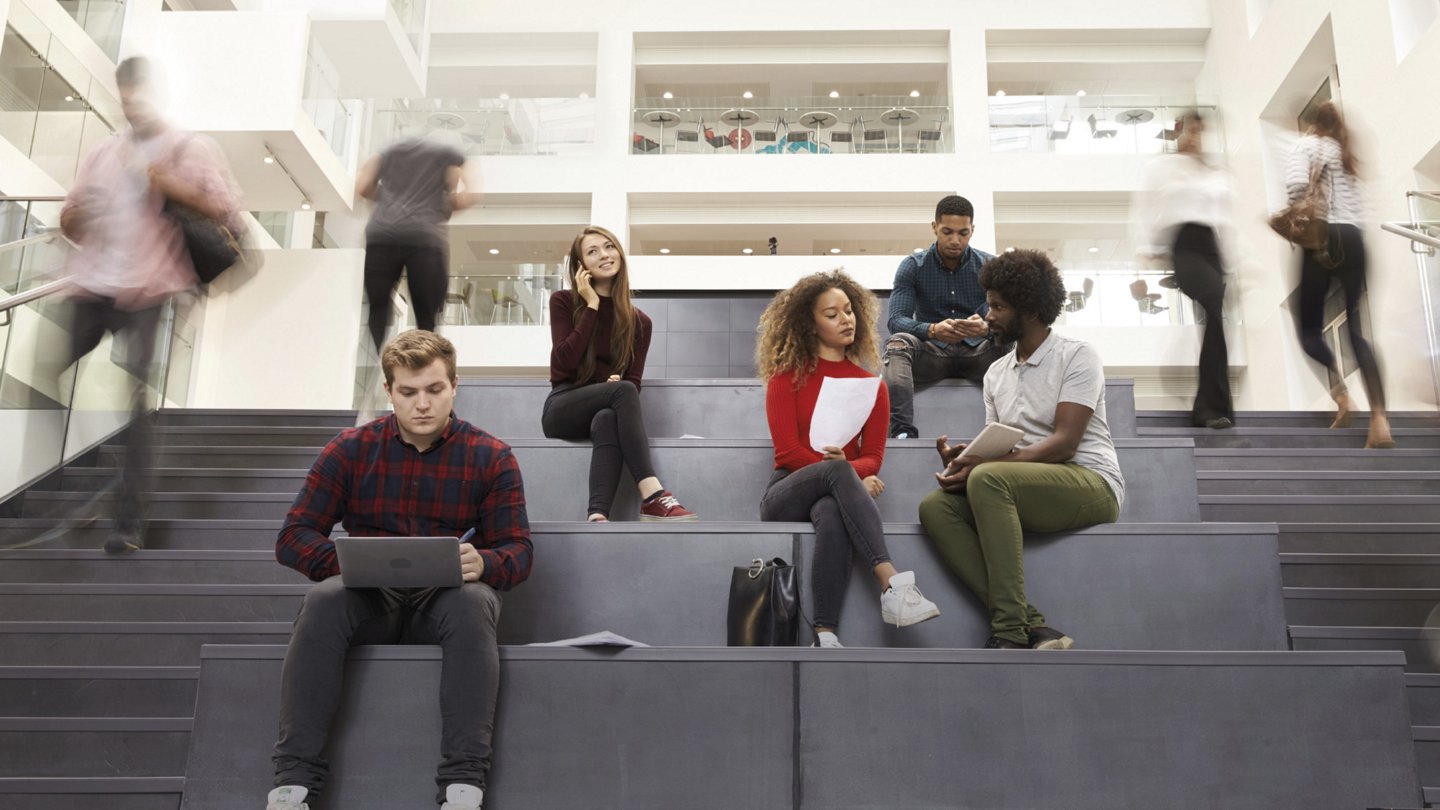 Gruppe von Studierenden sitzt auf Treppen in einem modernen Gebäude und unterhält sich, während andere Personen die Treppen hinauf- und hinuntergehen.