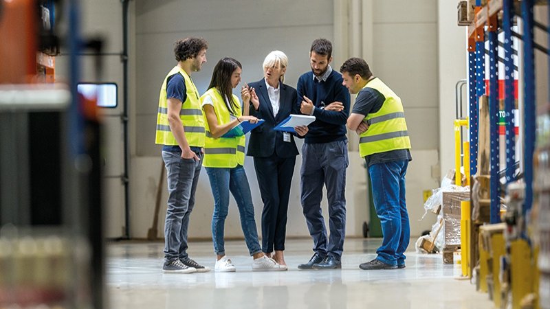 eine Gruppe von Menschen, teils in Schutzwesten, teils in Anzügen steht beiienander und blickt auf eine Frau mit einer Mappe in der Hand, sie gestikuliert und scheint etwas zu erklären