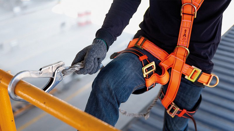 um eine Hüfte geschlungener Gurt, die Hand mit Handschuh hält ein Seil, das mit einem Karabiner um eine Stange gesichert ist.