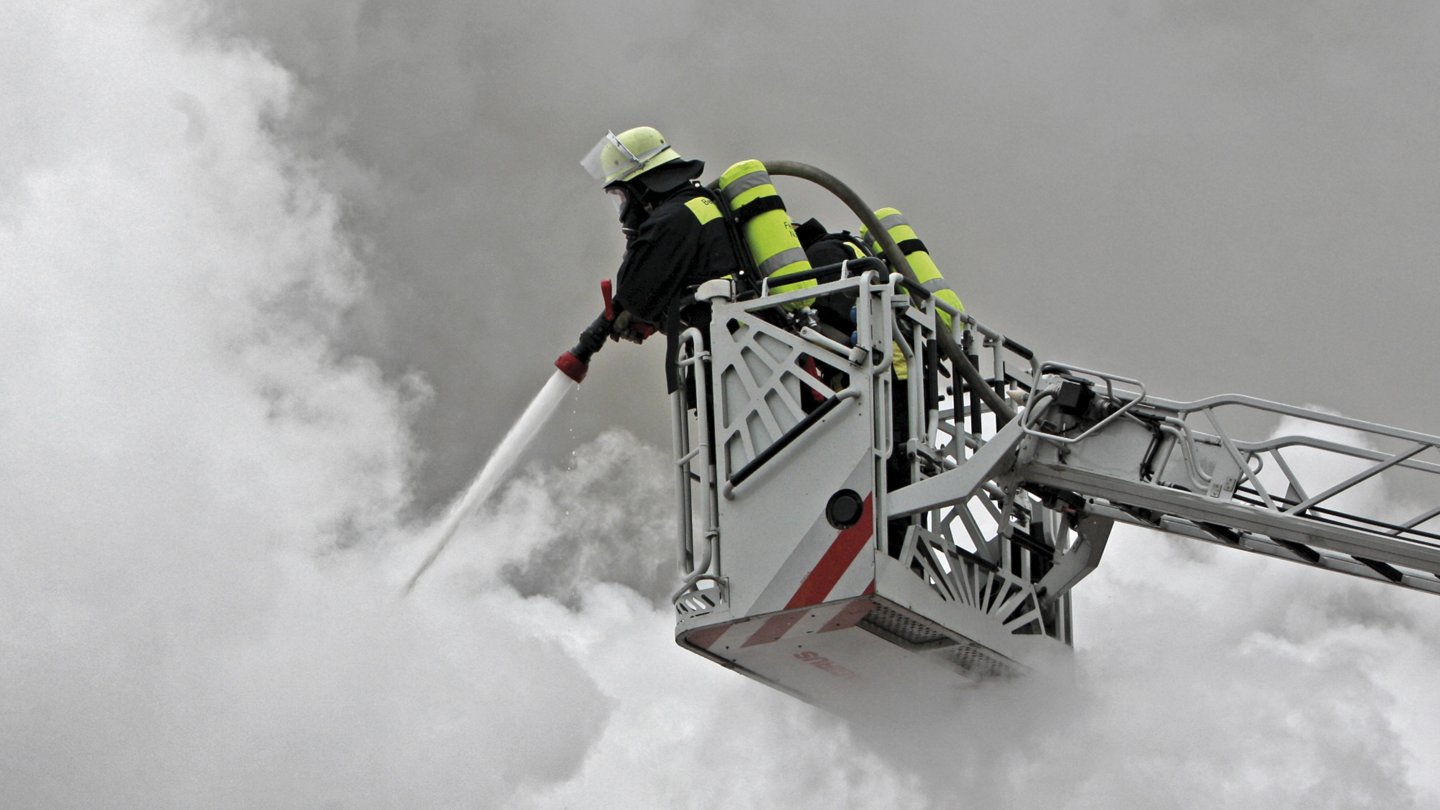 Ein Feuerwehrmann in Schutzkleidung und Atemgerät steht auf einer erhöhten Plattform eines Feuerwehrwagens und löscht mit einem Wasserschlauch dichten weißen Rauch. Der Feuerwehrmann arbeitet in großer Höhe, um einen schwer zugänglichen Brandherd zu bekämpfen.