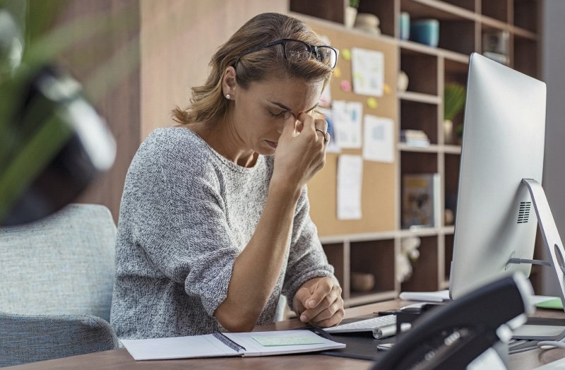 Frau sitzt an einem Schreibtisch vor einem Computer, hält sich die Stirn und wirkt gestresst oder nachdenklich.