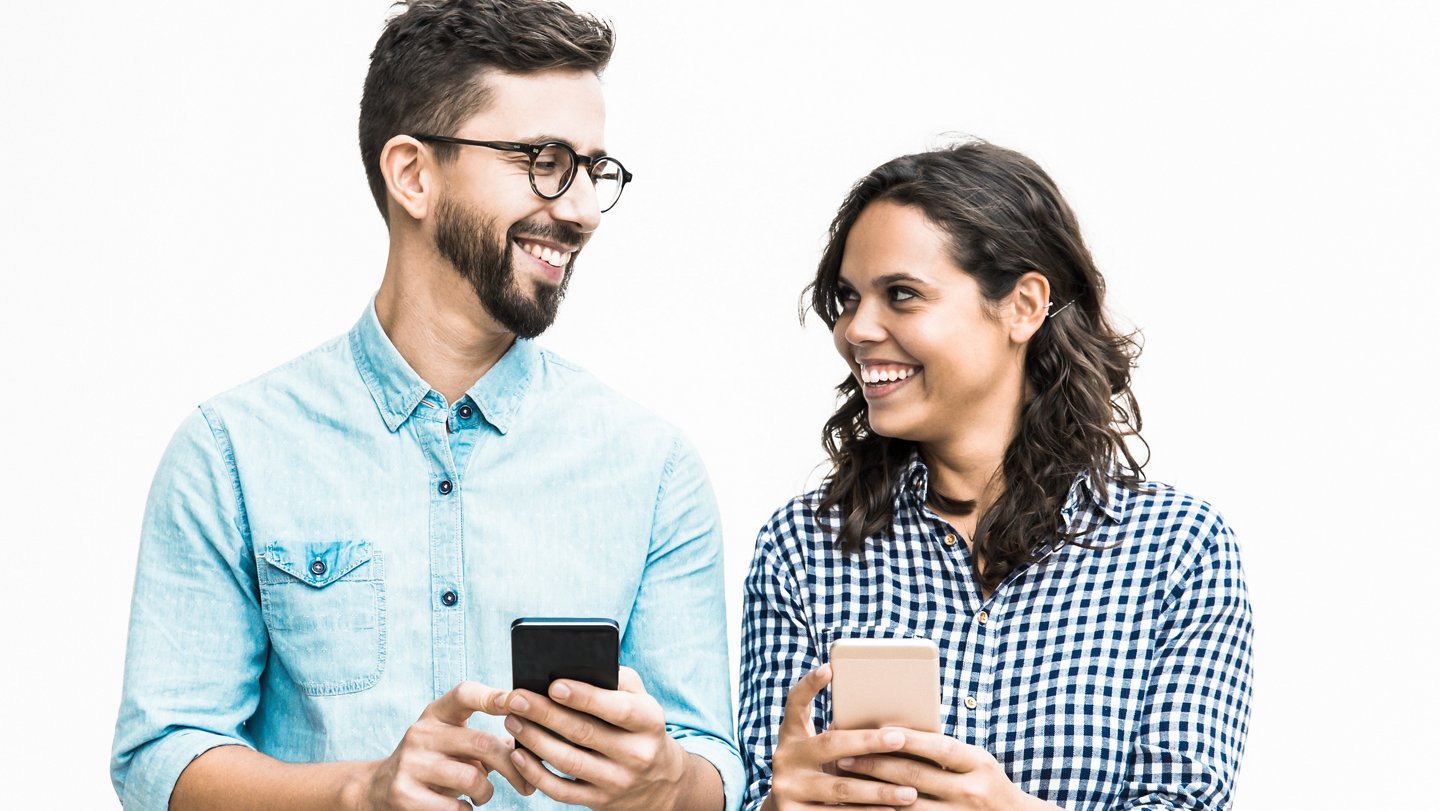 Happy cheerful couple with cellphones chatting and laughing. Young woman in casual and man in glasses posing isolated over white background. Good news concept