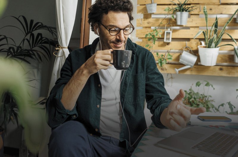 Ein Mann mit Brille und Kopfhörern hält eine Tasse in der Hand und lächelt. Er sitzt in einem Raum mit vielen Pflanzen, darunter einige in hängenden Töpfen und auf einem vertikalen Holzregal arrangiert. Ein Vorhang und ein Fensterrahmen sind im Hintergrund sichtbar.