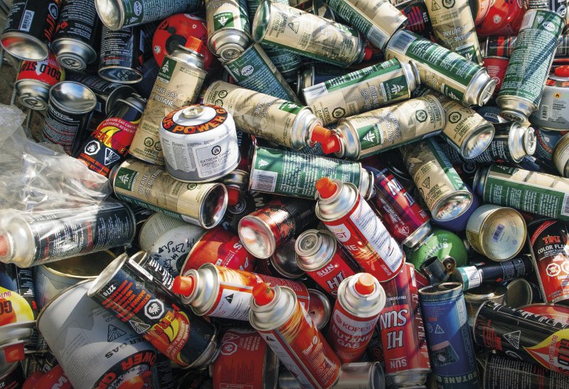 Ein Metallkorb, der mit leeren Spraydosen und Gaskartuschen gefüllt ist. Die Dosen sind bunt gemischt und scheinen für die Entsorgung oder das Recycling vorbereitet zu sein. Die Kartuschen stammen von verschiedenen Produkten, wie Brennstoff und Farben, die teils Gefahrensymbole tragen.