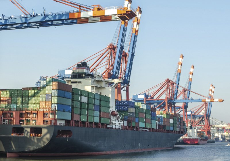 Ein großes Frachtschiff, beladen mit Containern in verschiedenen Farben, liegt in einem Hafen. Über dem Schiff ragen mehrere blaue und orangefarbene Containerkräne empor, die für das Be- und Entladen der Fracht verwendet werden. Im Hintergrund sind weitere Schiffe und Kräne zu sehen, während der klare Himmel das maritime Hafenbild abrundet.