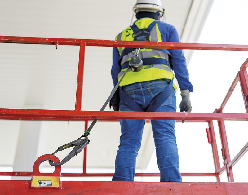 ein Arbeiter in Schutzkleidung und mit Helm in Rückenansicht, gesichert über einen Brustgurt mit einem großen Karabiner an der Bühne