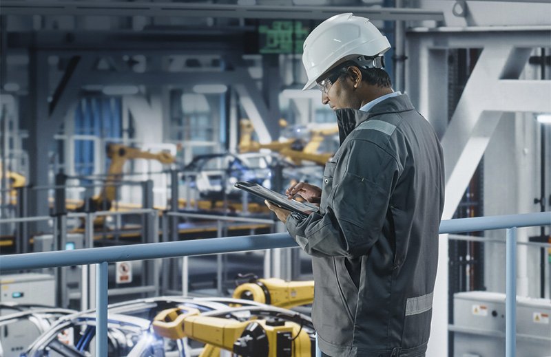 ein Mann mit Arbeitskleidung mit einem Tablet beobachtet Roboterarme, die Autos zusammenbauen