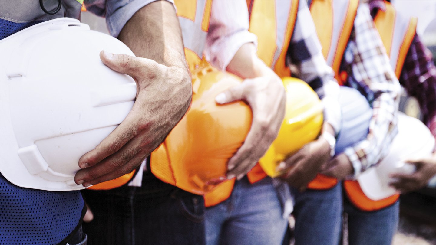 mehrere hintereinander stehende Arbeiter, jeder von ihnen hält einen Schutzhelm in der Hand