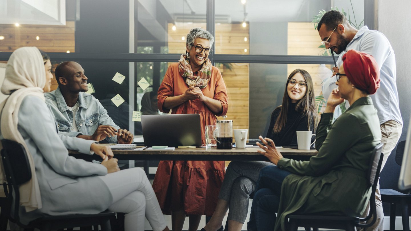 Eine diverse Gruppe von Menschen sitzt lachend und diskutierend um einen Tisch in einem modernen Büro.