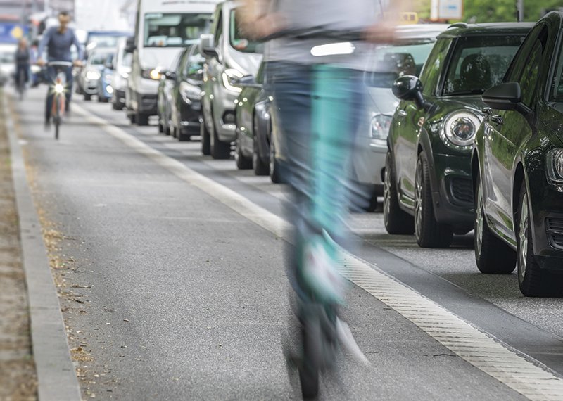 Straße mit vielen Autos, daneben überholen Fahrradfahrer und Scooter