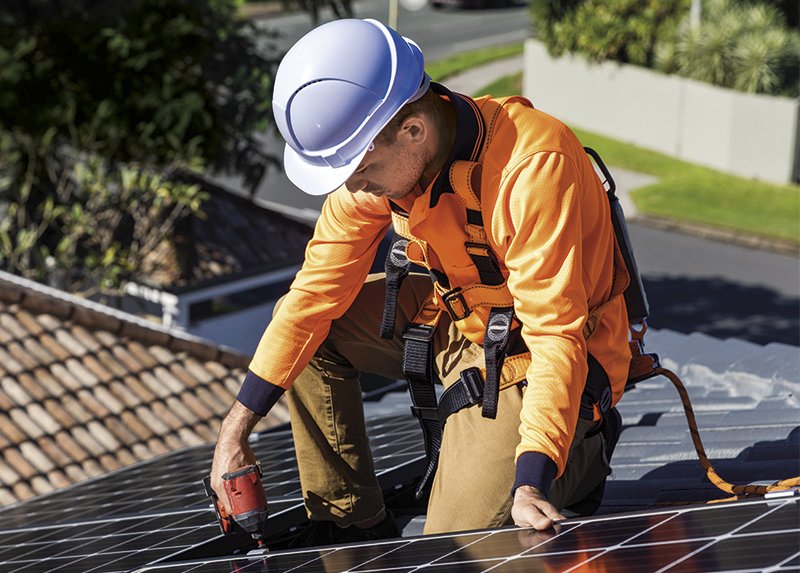 Solar panel technician with drill installing solar panels on house roof on a sunny day.