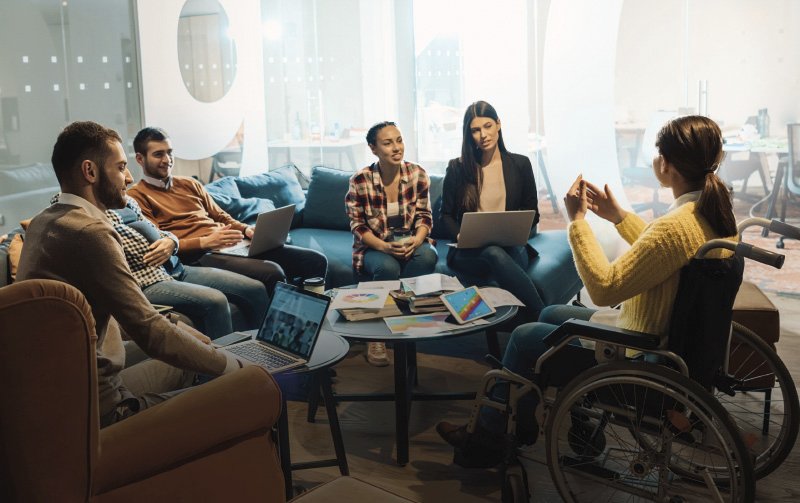 Eine Gruppe junger Menschen, darunter eine Person im Rollstuhl, diskutiert in einem modernen Büro mit Laptops und Notizen auf einem Tisch.