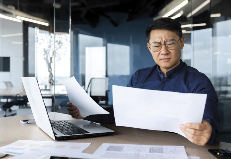 Ein Mann mit Brille sitzt in einem modernen Büro vor einem Laptop und hält zwei Blätter Papier in den Händen. Er blickt konzentriert auf die Dokumente, die auf dem Schreibtisch verteilt sind. Im Hintergrund ist eine Glaswand zu sehen, durch die ein helles Büro erkennbar ist.