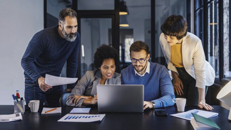 Vier Personen sitzen und stehen an einem Tisch in einem modernen Büro und schauen gemeinsam auf einen Laptop. Eine Frau zeigt mit dem Finger auf den Bildschirm, während ein Mann neben ihr aufmerksam schaut. Eine weitere Frau und ein Mann beugen sich interessiert über die Szene. Auf dem Tisch liegen Papiere, ein Stift und eine Kaffeetasse.