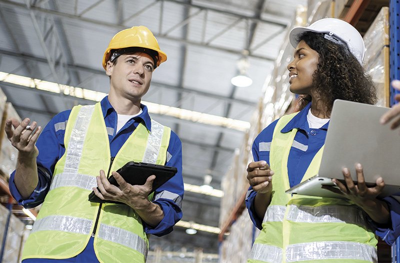 Arbeiter:innen mit Helm und Schutzweste stehen in einer Lagerhalle beisammen