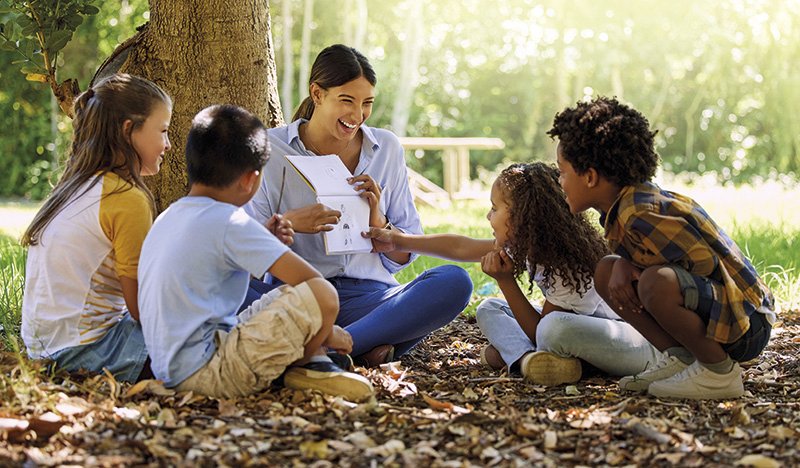 eine Frau und Kinder sitzen im Wald am Boden und besprechen eine Zeichnung, die die Lehrerin hält