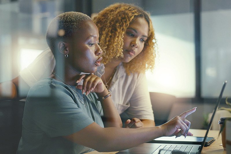 zwei Frauen sitzen vor einem Computer, eine der beiden zeigt auf den Monitor des Notebooks