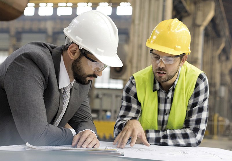 Zwei Männer mit Schutzhelm und Schutzweste stehen bei einem Schreibtisch beisammen und besprechen einen Plan