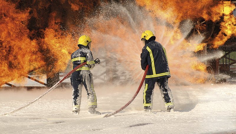 Feuerwehrleute in einem Löschschaum vor Flammen.
