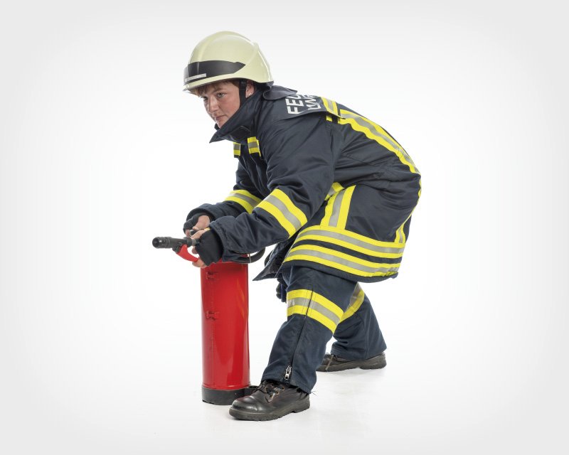 Ein Feuerwehrmann in voller Schutzkleidung, inklusive Helm, kniet nieder und hält einen Feuerlöscher, bereit, diesen zu benutzen. Der Hintergrund ist weiß, wodurch der Fokus vollständig auf den Feuerwehrmann und den Feuerlöscher gelenkt wird.