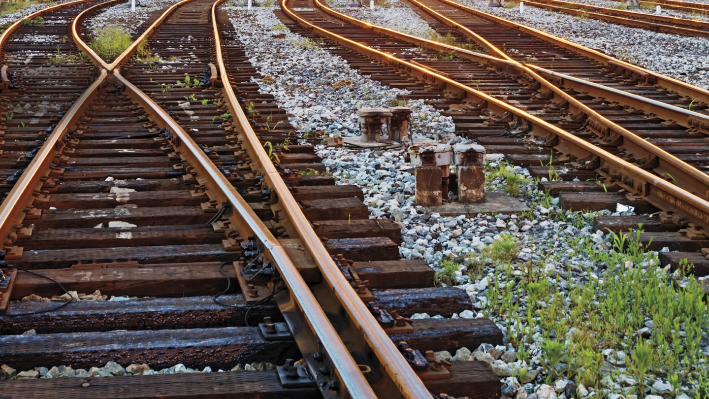 Ein weitläufiges Bahngelände mit mehreren Gleisen, die sich verzweigen und in verschiedene Richtungen verlaufen. Im Hintergrund sind zahlreiche Güterwaggons und Eisenbahnsignale zu sehen. Über den Gleisen verläuft eine Linie von Beleuchtungen, die das Gelände in warmes Licht tauchen. Der Himmel ist klar und strahlt in einem hellen Blau, was auf einen ruhigen, frühen Morgen oder späten Abend hinweist.