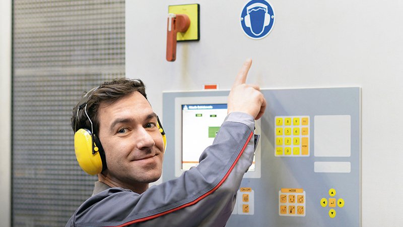ein Mann mit Gehörschutz steht in einem Maschinenraum und deutet auf das Symbol für das Tragen von Gehörschutz