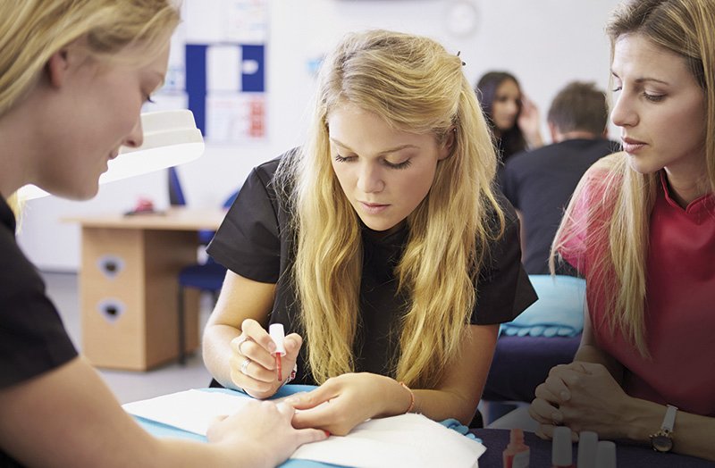 eine junge Nageldesignerin lackiert den Nagel einer Frau, eine andere Frau blickt ihr freundlich prüfend über die Schulter