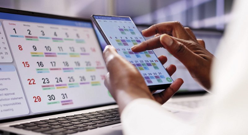 Eine Person hält ein Smartphone in der Hand, auf dem ein digitaler Kalender angezeigt wird, und bedient es. Im Hintergrund sind die Bildschirme von Laptops zu sehen, die ebenfalls Kalenderansichten zeigen.