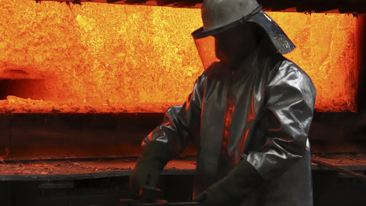 Im Hintergrund der Hochofen mit orange-gelbem Feuer, im Vordergrund eine dunkle Arbeiter-Silhouette