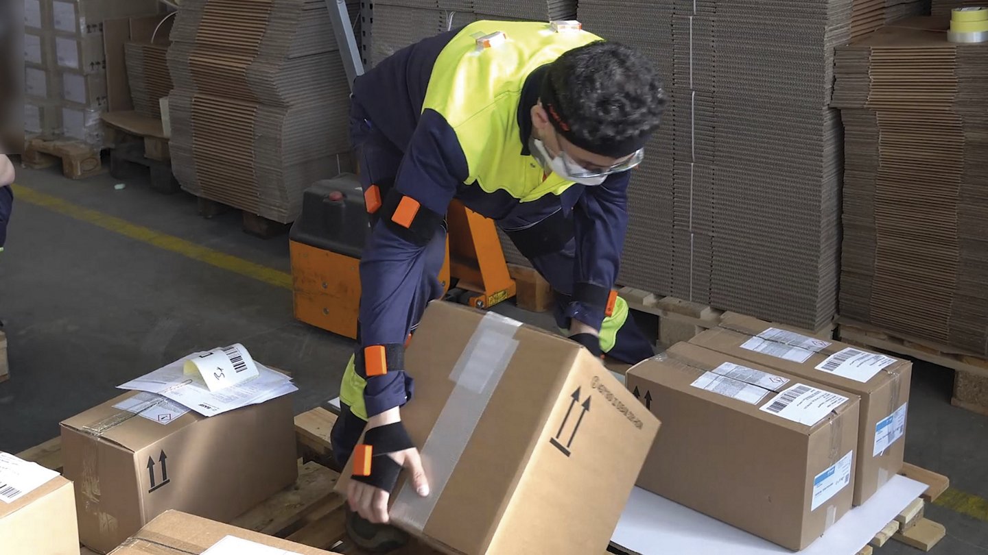 Ein Arbeiter in Schutzkleidung und mit Schutzbrille hebt einen Karton in einem Lagerbereich an. Im Hintergrund sind gestapelte Kartons und Verpackungsmaterial zu sehen. Der Arbeiter verwendet orangefarbene Sensoren an Armen und Beinen, möglicherweise zur Überwachung seiner Bewegungen oder Haltung. Eine zweite Person, deren Gesicht unscharf ist, sitzt links und überprüft Dokumente oder Geräte.